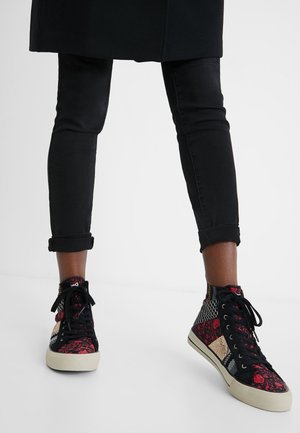 Nogi w czarnych, podwiniętych obcisłych dżinsach i wysokich trampkach z czerwono-czarn-beżowym wzorem patchworkowym na białej podłodze.