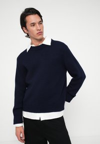 Nuori mies, joka on pukeutunut tummansiniseen neuleeseen valkoisen paidan päällä, seisoo yksi käsi takanaan ja katsoo vasemmalle yksinkertaista valkoista taustaa vasten.