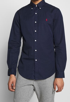 Mężczyzna ubrany w granatową koszulę na guziki z czerwonym wyhaftowanym logo na piersi oraz jasnoszare spodnie.