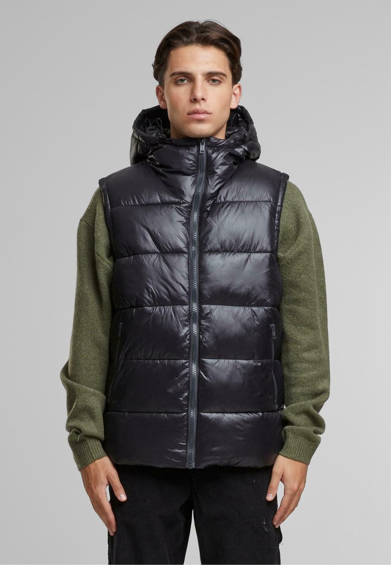 Polstret svart vest med glidelås og hette, med et quiltet design og blank tekstur. Båret over en grønn langermet skjorte.