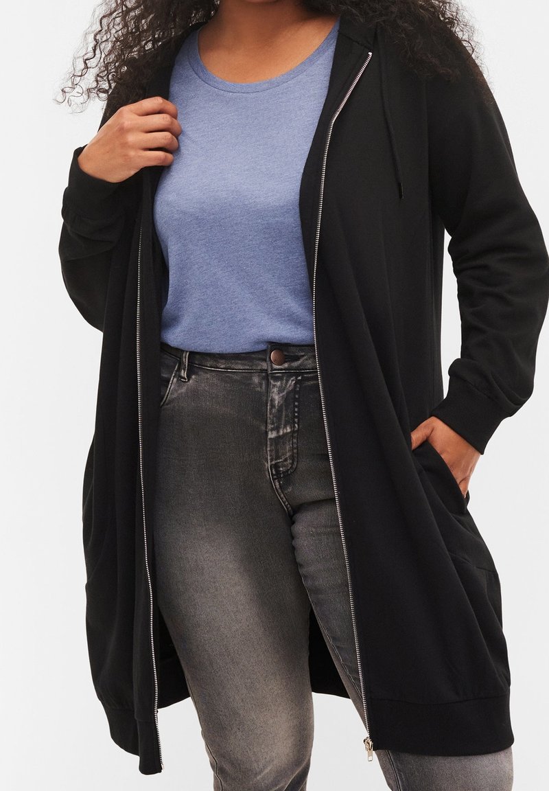 Cárdigan largo negro con cremallera frontal, dos bolsillos laterales y capucha, usado sobre una camiseta azul y pantalones gris oscuro. Textura de tela suave.