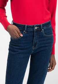 Jeans de mezclilla azul oscuro con una cintura alta, con un diseño de cinco bolsillos, costuras claras y cierre de botón metálico. Mano en el bolsillo.