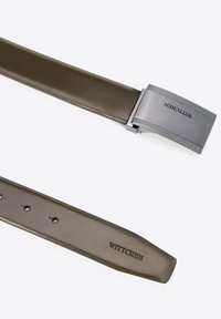 Bruine leren riem met een metalen speld. De speld heeft een gestructureerd oppervlak en de merknaam "WITTCHEN" is gegraveerd.