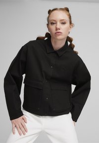 Veste noire avec un col, boutons-pression, deux poches devant et tissu texturé ; portée sur un pantalon blanc. Torse détaché exposé.