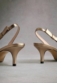 Gold metallic heeled sandals with slim straps and bows. The shoes have a tapered stiletto heel and a smooth, shiny finish.