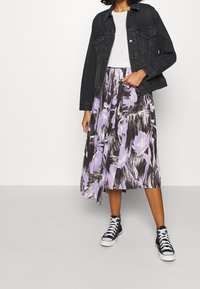 Svart denimjacka över en vit t-shirt, kombinerad med en knälång, blommönstrad kjol i lila och svart, samt svarta högskaftade sneakers.