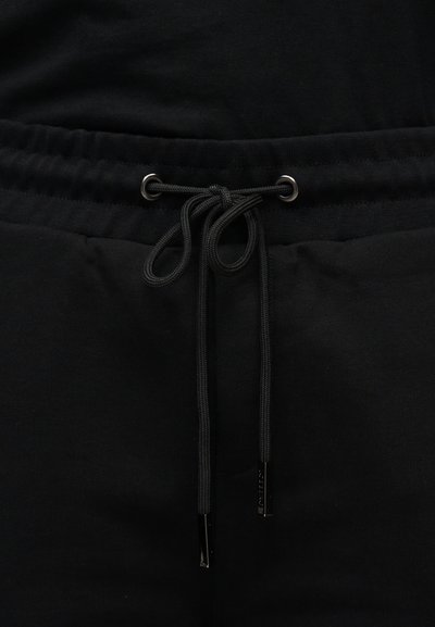 Gros plan sur la ceinture d'un pantalon de survêtement noir avec un élastique et un cordon noir noué passant à travers des œillets métalliques.