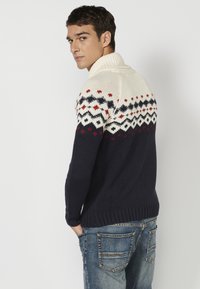 Suéter de punto con cuello alto, sección superior color crema con patrones geométricos en rojo y marino, y cuerpo en marino oscuro. No se observan otros detalles.