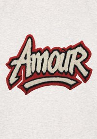 Patch brodé portant le mot "Amour", avec des contours rouge vif et noir sur un fond crème, et une finition en point texturé.