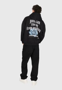 Uomo che indossa un hoodie nero e pantaloni, sneakers bianche, di spalle rispetto alla fotocamera, il testo sull'hoodie recita "puoi rialzarti da qualsiasi cosa" con una grafica di una rosa blu.