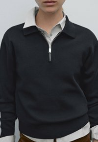 Svart stickad tröja med dragkedja fram, med krage och en vit skjorta under. Strukturerat tyg med ribbade muddar och nederkant.