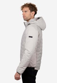 Chaqueta acolchada gris claro con un patrón acolchado, capucha y distintivo negro en la manga; presenta una textura suave y diseño ajustado.