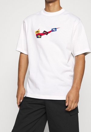T-shirt blanc avec des logos "Jump Man" colorés reliés par un trait rouge, porté avec un pantalon cargo noir, torse et bras visibles.