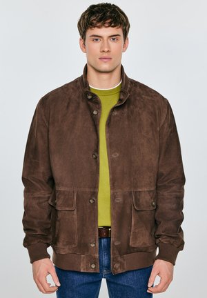 Brązowa zamszowa kurtka ze stójką, czterema przednimi kieszeniami, zapinana na guziki, z dzianinowymi mankietami i gładką fakturą, noszona na zielonej koszuli.