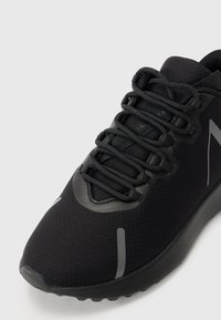 Zapatilla deportiva negra con tejido texturizado, cordones gruesos negros y franjas reflectantes grises en la punta y el costado, exhibida sobre un fondo blanco.