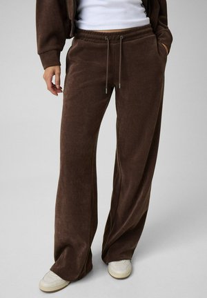 Personne portant un pantalon large en velours côtelé marron avec une taille à cordon, un haut blanc et des baskets blanches.