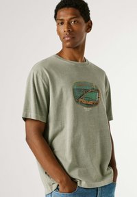 Noor mees, kes kannab rohelist Ramsgate Lido graafilist t-särki ja siniseid teksaseid, seisab käed taskutes lihtsa tausta ees.