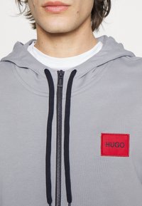 Sudadera con capucha gris que presenta una cremallera negra, cordones y un parche de logo bordado en rojo. Material de mezcla de algodón con textura acanalada.