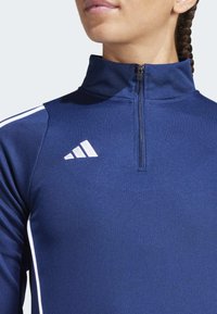 Jūras spēku sporta krekls ar puspagaru rāvējslēdzēju, izgatavots no gludas auduma. Iezīmēts ar baltām svītrām pāri pleciem un Adidas logotipu krūtīs.