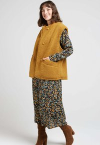 Gilet sans manches en fausse fourrure de couleur moutarde avec un col arrondi, deux poches avant, porté sur une robe à motifs floraux, associé à des bottes marron.