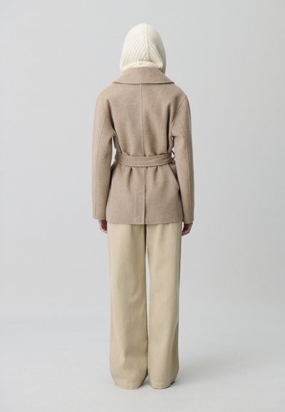 Personne de dos, portant un pantalon large beige, un manteau camel ceinturé et un capuchon en tricot crème, sur un fond uni.