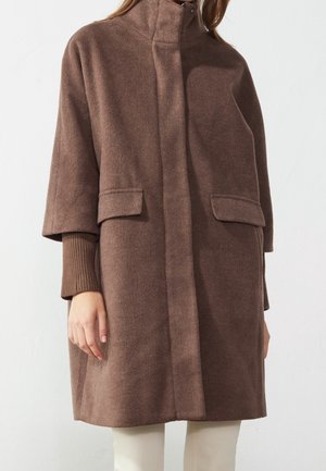 Vrouw die een knielange bruine wollen jas draagt met verborgen knopen aan de voorkant en klepzakken, gecombineerd met crèmekleurige broek en een bruine trui.