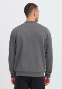 Sweatshirt gris avec col ras du cou, manches longues, poignets en côte et ourlet. Texture lisse, design minimal, porté par une personne de dos.