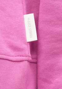 Ύφασμα ροζ φούτερ με λεία υφή. Διαθέτει λευκή ετικέτα με εκτύπωση "J.JK STUDIO". Ορατές ραφές και ριμπ μανσέτες.