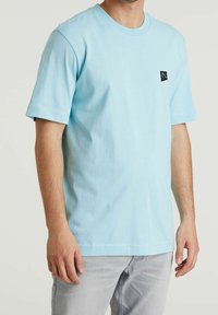 Hellblaues Baumwoll-T-Shirt mit kurzen Ärmeln und Rundhalsausschnitt. Verfügt über einen kleinen schwarzen Logo-Patch auf der linken Brust. Einfaches Design.