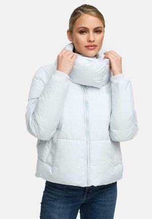Vrouw die de hoge kraag van een lichtblauwe gewatteerde jas aanpast, draagt donkere jeans, tegen een effen witte achtergrond.