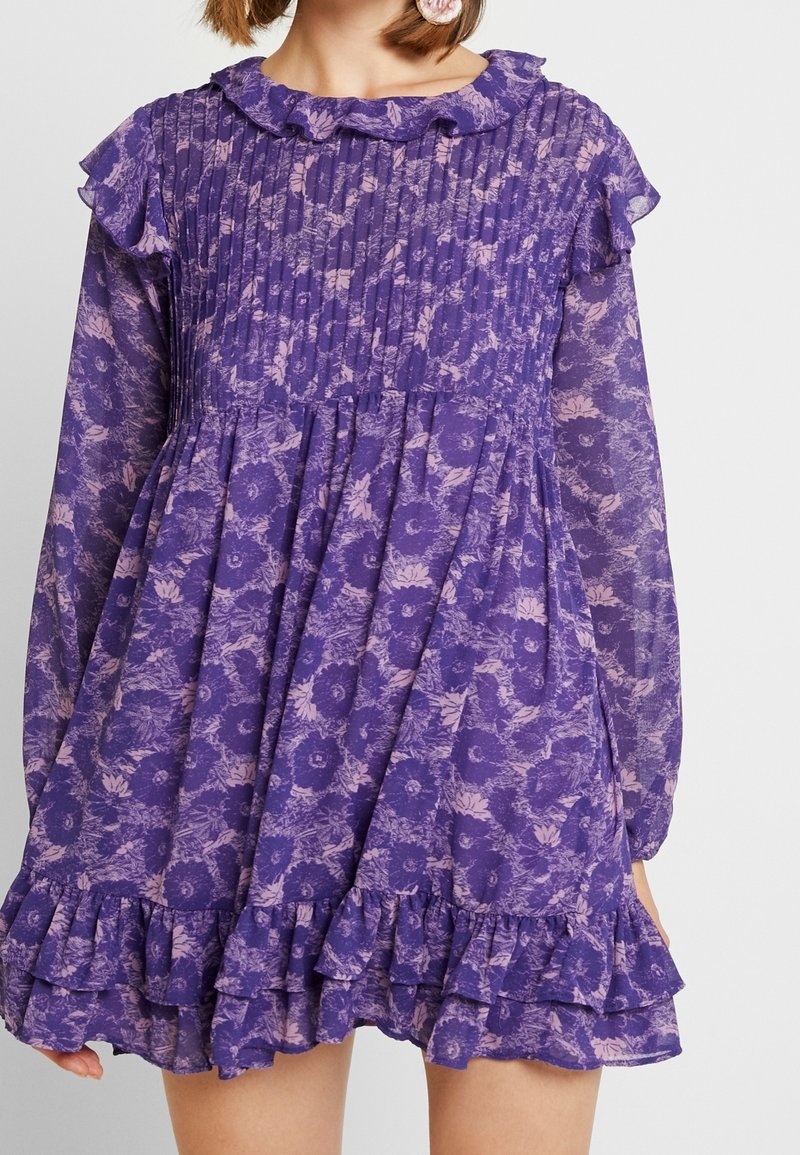 Robe florale violette avec manches longues en voile transparent, col et ourlet à volants, et corsage plissé portée par une personne debout devant un fond uni.