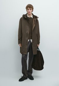 Brązowy wodoodporny płaszcz z kapturem, noszony na brązowym swetrze i białej koszuli. Szare spodnie i czarna torba na ramię dopełniają stylizację.