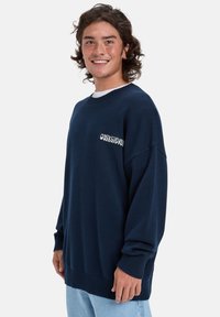 Sweater azul marinho com punhos e barra canelados, apresentando um logótipo branco no peito. Feito de um tecido macio e texturizado com um corte descontraído.