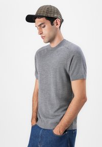 Γκρι κοντομάνικο πουλόβερ με στρογγυλή λαιμόκοψη και ριχτό στρίφωμα, σε συνδυασμό με καφέ καπέλο με καρό σχέδιο που έχει μαύρο γείσο.