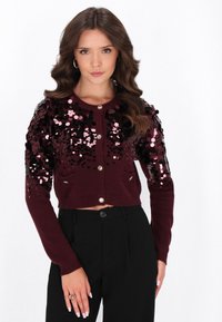Frau mit welligem braunem Haar, die eine burgunderrote, kurz geschnittene Strickjacke mit pinken und schwarzen Pailletten, schwarze hochgeschnittene Hosen und goldene Creolen trägt.
