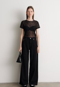 Camiseta preta em malha transparente com riscas horizontais, combinada com calças largas de cintura alta pretas. Os acessórios incluem uma pequena mala preta.