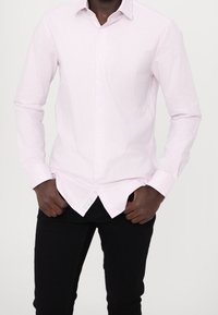Homme portant une chemise à rayures verticales rose clair et blanche avec des boutons et un pantalon noir, debout les mains dans les poches.