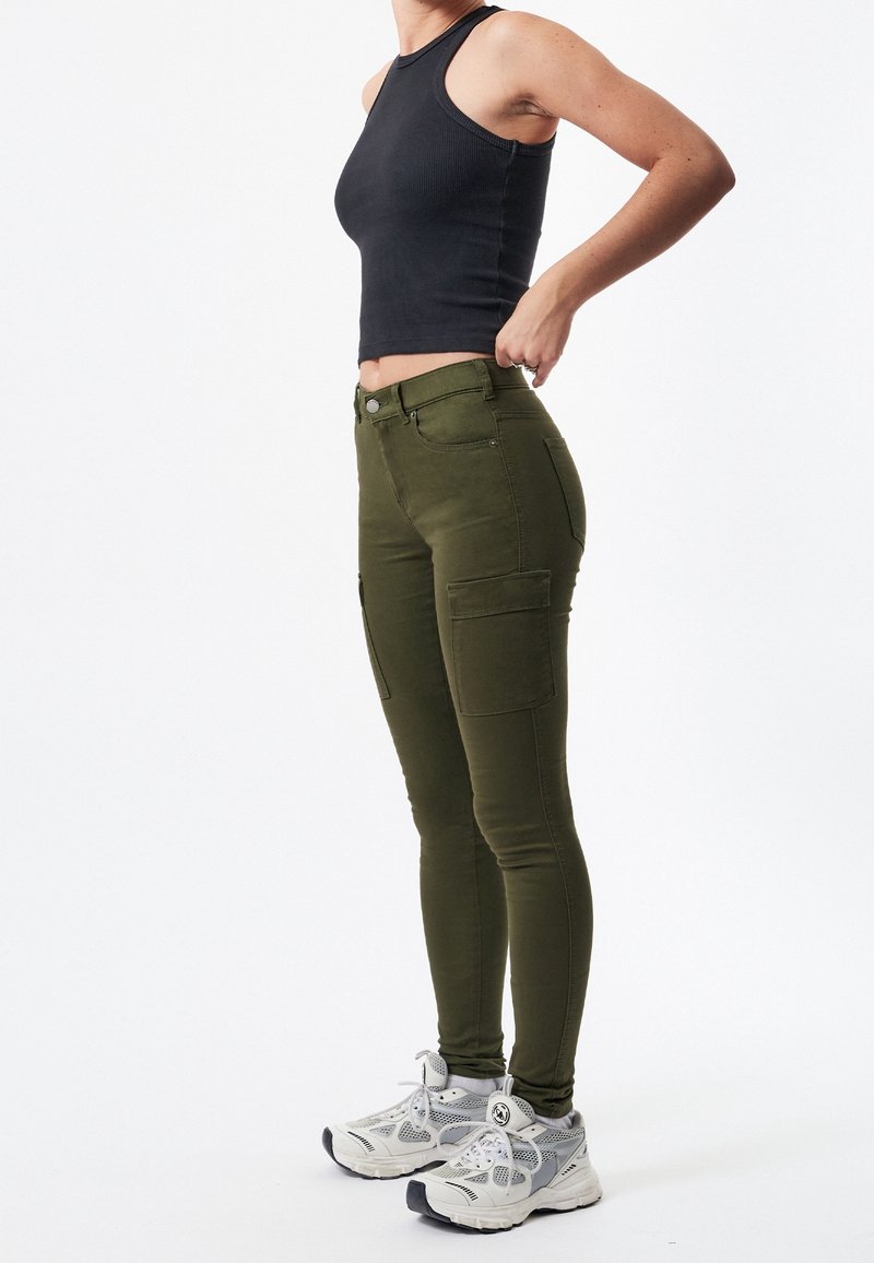 Personne portant un pantalon cargo vert olive, un haut sans manches noir et des baskets blanches, debout devant un fond blanc uni.
