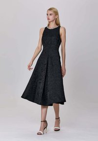 Femme portant une robe midi sans manches noire texturée avec une jupe évasée et un col orné, associée à des sandales à talons hauts noir et blanc.