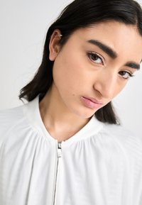 White zip-up jacket with gathered fabric at the neck, set against a neutral background. The model's hair is dark and straight, with an earring visible.