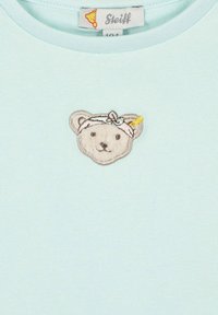 Top de algodón azul claro con un rostro de oso bordado, un lazo y un acento amarillo. Incluye una etiqueta con la marca "Steiff".