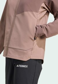 Veste Terrex avec un design bicolore en marron clair et marron foncé, fermeture zippée et poches latérales, associée à un pantalon sombre affichant un logo.