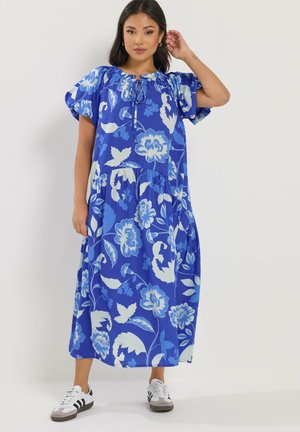 Kobieta ubrana w niebieską sukienkę midi w kwiaty z bufkami i białe trampki, stojąca na jednolitym jasnym tle.