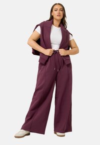 Weinroter, übergroßer Weste über einem weißen T-Shirt, kombiniert mit locker sitzenden, weinroten Hosen mit elastischem Kordelzugbund und weicher Textur.