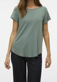 Top de manga corta en tejido verde apagado, con cuello redondo, forma drapeada y un dobladillo ligeramente curvado. Combinado con jeans oscuros.