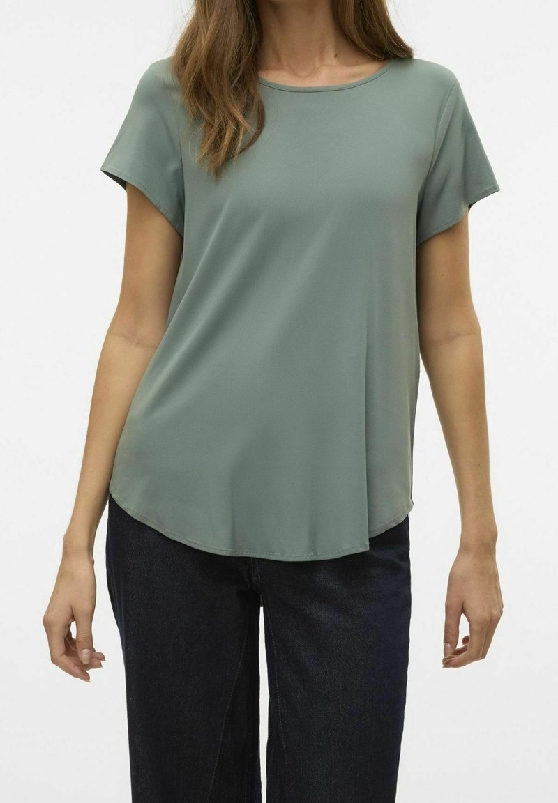 Top de manga corta en tejido verde apagado, con cuello redondo, forma drapeada y un dobladillo ligeramente curvado. Combinado con jeans oscuros.