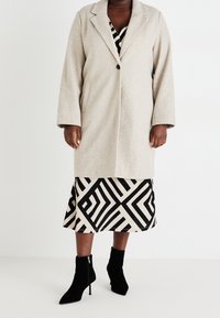 Beżowy płaszcz z mieszanki wełny z kołnierzem z klapami i pojedynczym zapięciem na guzik, noszony na spódnicy z geometrycznym wzorem oraz czarnych botkach za kostkę.