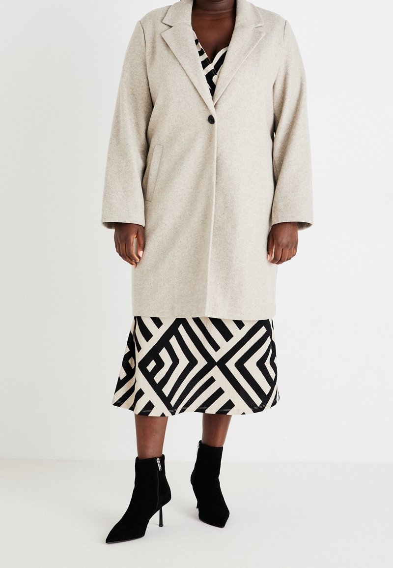 Beżowy płaszcz z mieszanki wełny z kołnierzem z klapami i pojedynczym zapięciem na guzik, noszony na spódnicy z geometrycznym wzorem oraz czarnych botkach za kostkę.