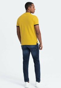 Gul polo-shirt med sort ribbet krave, parret med mørkeblå jeans og hvide sneakers. Skjorten har sideslids ved kanten.