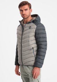 Vuoritettu hupullinen takki harmaana ja tummanharmaana, jossa on vaakasuora tikattu muotoilu, vetoketjulliset sivutaskut ja logo rinnassa.